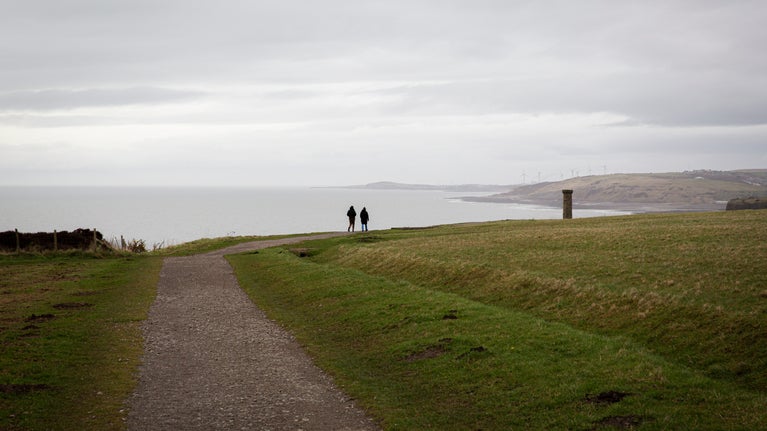 Walkers follow a coastal path along the cliffs at Whitehaven on a brisk day in winter, facing towards a grey sea and an overcast sky.